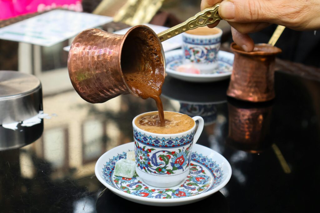 A hand pours Turkish coffee from a copper cezve into a decorative cup. Outdoor ambiance.
